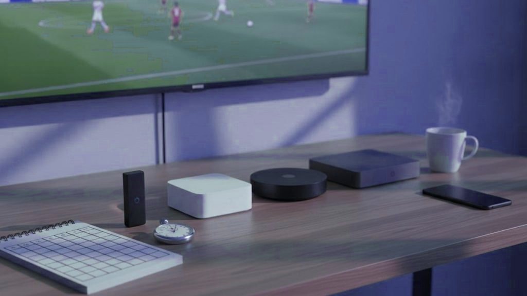 Five streaming devices, a notebook scoring grid and a stopwatch in front of a TV showing a football match